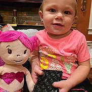 Genesis is registered to the contest to win money with this photo: toddler, child, plush_toy, mermaid_doll, pink_hair, glitter, wallet, indoor, brick_wall, earrings, short_hair, pink_shirt, black_skirt, toy, person, cute, smile, sitting, holding, cozy
