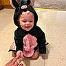 Genesis is registered to the contest to win money with this photo: toddler, child, costume, bat_costume, indoor, floor, tile_floor, carpet, hand_holding, smiling, cute, baby, person, cabinets, playful, kneeling, pink, black, ears, happy