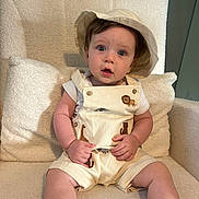 Freddie is registered to the contest to win money with this photo: baby, child, hat, overalls, animal_embroidery, armchair, cushion, plush_fabric, curious_expression, sitting, indoor, cream_color, cute, portrait, infant, clothing, face, hands, legs, young_child