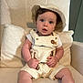 baby, child, hat, overalls, animal_embroidery, armchair, cushion, plush_fabric, curious_expression, sitting, indoor, cream_color, cute, portrait, infant, clothing, face, hands, legs, young_child