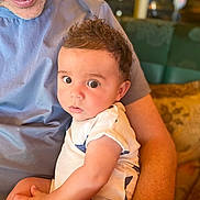 Joseph joined the competition — help win amazing prizes! baby, child, infant, person, adult, arm, curly_hair, blue_shirt, white_onesie, indoor, couch, cushion, skin, face, eyes, hand, portrait, closeup, comfort, warm_lighting
