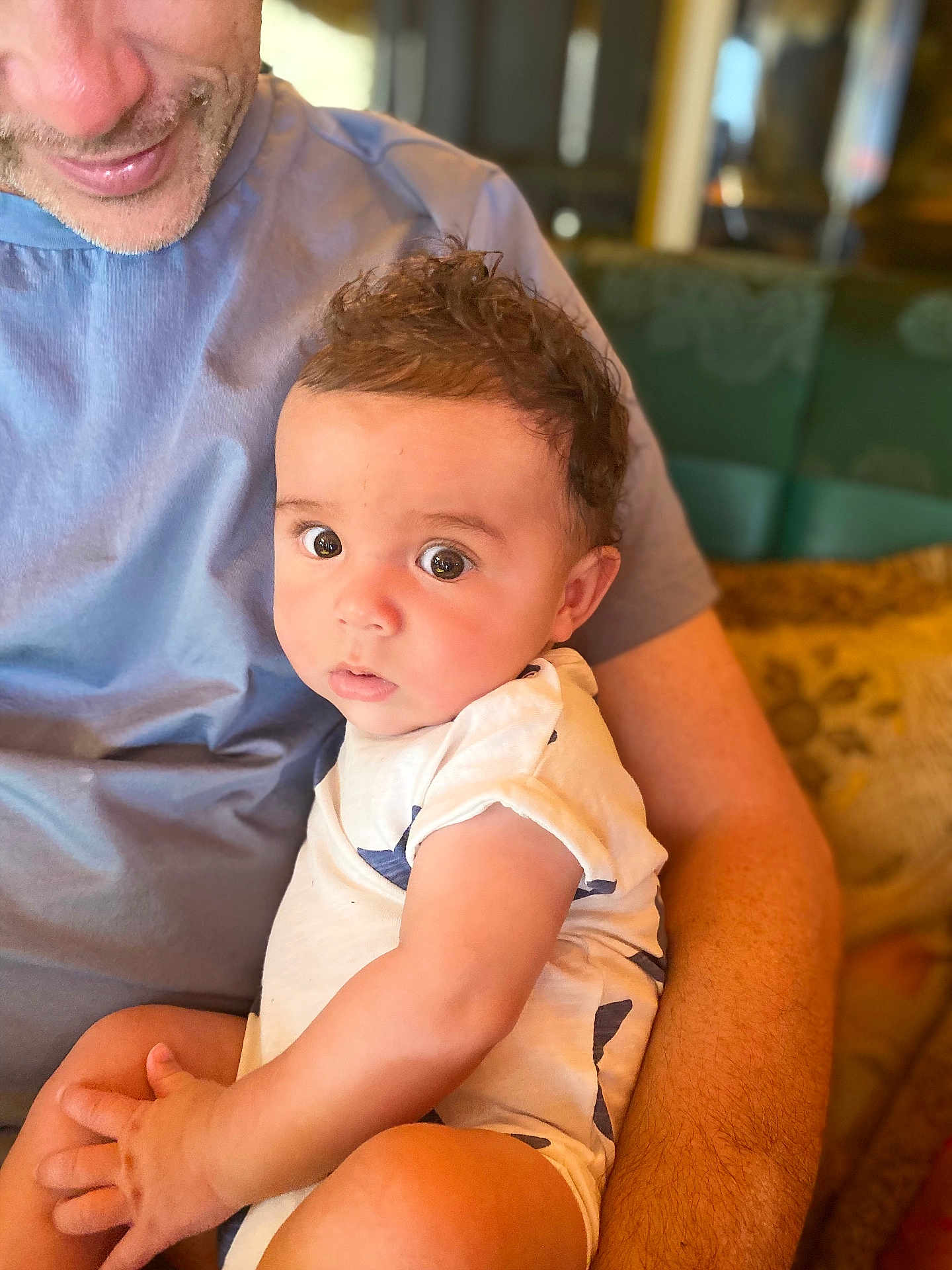 Joseph joined the competition — help win amazing prizes! baby, child, infant, person, adult, arm, curly_hair, blue_shirt, white_onesie, indoor, couch, cushion, skin, face, eyes, hand, portrait, closeup, comfort, warm_lighting