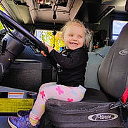 Helen is registered to the contest to win money with this photo: child, toddler, smile, steering_wheel, vehicle_interior, seat, clothing, shoes, window, autumn, tree, dashboard, seatbelt, happy, person, indoor, playful, cute, vehicle, young_child