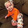 baby, child, smiling, orange_clothing, headband, bow, pumpkin_pattern, pants, sitting, couch, brown_furniture, foot, hand, face, blue_eyes, happy, indoors, cute, infant, portrait