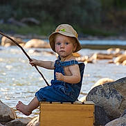 Finn is registered to the contest to win money with this photo: toddler, child, river, water, rocks, fishing, stick, hat, overalls, yellow_boots, outdoor, nature, summer, sunlight, wooden_box, barefoot, portrait, playful, candid, adventure