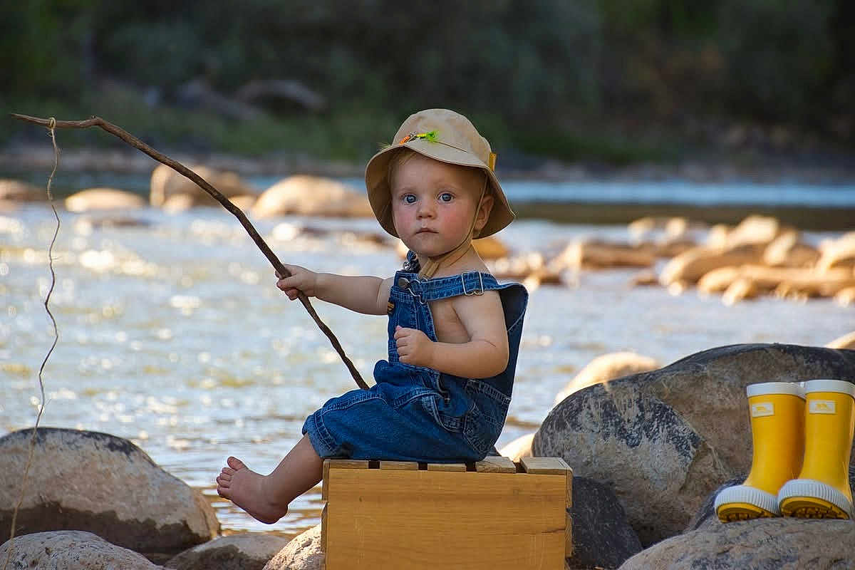 Finn is registered to the contest to win money with this photo: toddler, child, river, water, rocks, fishing, stick, hat, overalls, yellow_boots, outdoor, nature, summer, sunlight, wooden_box, barefoot, portrait, playful, candid, adventure