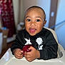 apple, baby, black_sweatshirt, child, cute, expression, face, food, hands, happy, high_chair, home, indoor, person, portrait, seated, smiling, table, toddler, young_child