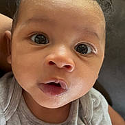 Isaiah joined the competition — help win amazing prizes! baby, infant, face, eyes, mouth, curly_hair, grey_shirt, text_on_clothing, sitting, indoor, closeup, person, child, cute, expression, skin, head, young, portrait, curious