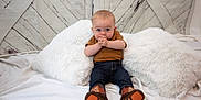 Trenton is registered to the contest to win money with this photo: baby, child, infant, blue_eyes, brown_shirt, jeans, boots, white_pillows, fluffy, white_sheet, wooden_wall, rustic, sitting, hands_clasped, curious, indoor, portrait, cute, young, playful
