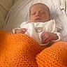 newborn, baby, sleeping, blanket, orange, knitted, crib, white_clothing, bear_embroidery, infant, hand, face, peaceful, cozy, soft, texture, portrait, indoors, resting, cute