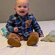 Ethan joined the competition — help win amazing prizes! baby, child, smiling, sitting, plaid_shirt, blue_shirt, brown_pants, indoor, bed, blanket, baby_wipes, happy, cute, portrait, infant, clothing, footwear, person, young_child, head