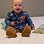 baby, child, smiling, sitting, plaid_shirt, blue_shirt, brown_pants, indoor, bed, blanket, baby_wipes, happy, cute, portrait, infant, clothing, footwear, person, young_child, head
