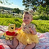 baby, child, tutu, headband, plush_toy, winnie_the_pooh, blanket, outdoor, grass, sunlight, greenery, tree, sky, cute, smile, sitting, playful, portrait, daylight, nature