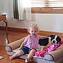 toddler, pacifier, dog, dog_bed, blanket, indoor, wooden_floor, child, curtains, window_blinds, pink_clothing, black_and_white_dog, hair, barefoot, furniture, relaxed, pet, home, comfort, floor