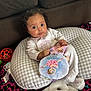 baby, big_eyes, boppy_pillow, checkered_pillow, child, colorful_carpet, couch, curly_hair, cute_expression, holding_toy, indoor, infant, onesie, patterned_blanket, pillow, playtime, plush_toy, sofa, stuffed_animal, toy_ball