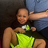 toddler, child, smiling, couch, neon_shorts, black_shirt, pen, happy, indoor, person, seated, relaxed, casual_clothing, shorts, bright_colors, face, cute, young_child, home, family