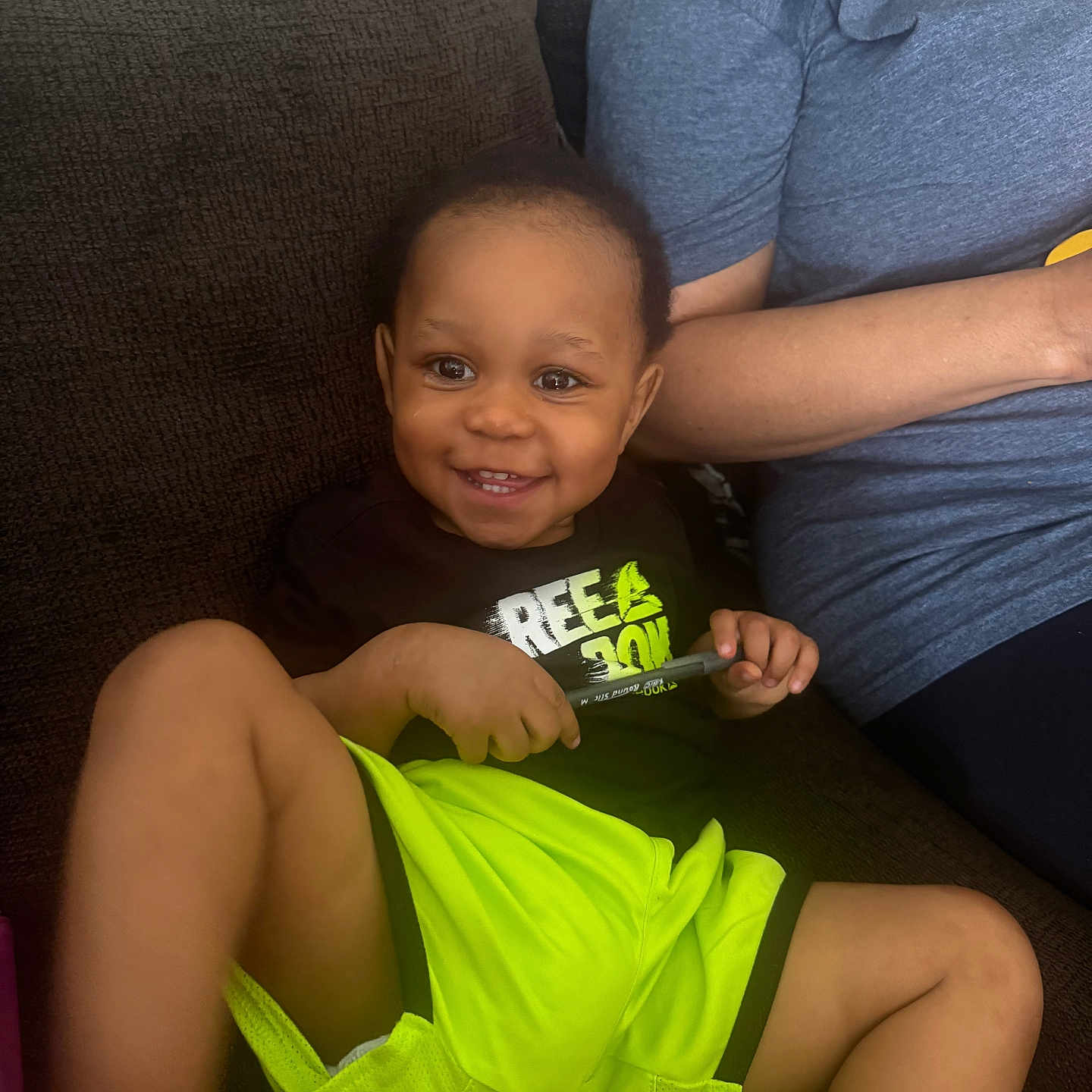 Jamari Holmes joined the competition — help win amazing prizes! toddler, child, smiling, couch, neon_shorts, black_shirt, pen, happy, indoor, person, seated, relaxed, casual_clothing, shorts, bright_colors, face, cute, young_child, home, family
