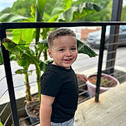 Dayven joined the competition — help win amazing prizes! toddler, child, smiling, black_shirt, gray_shorts, outdoor, railing, plants, greenery, concrete_floor, daylight, happy, portrait, short_hair, casual_clothing, nature, urban, pot, background, person