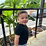 toddler, child, smiling, black_shirt, gray_shorts, outdoor, railing, plants, greenery, concrete_floor, daylight, happy, portrait, short_hair, casual_clothing, nature, urban, pot, background, person