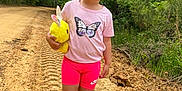 Skylar is registered to the contest to win money with this photo: child, girl, outdoor, dirt_path, greenery, stuffed_animal, yellow_bunny, purple_bow, pink_shorts, butterfly_tshirt, frozen_shoes, smiling, curly_hair, nature, trees, casual_clothing, footwear, summer, playful, walking
