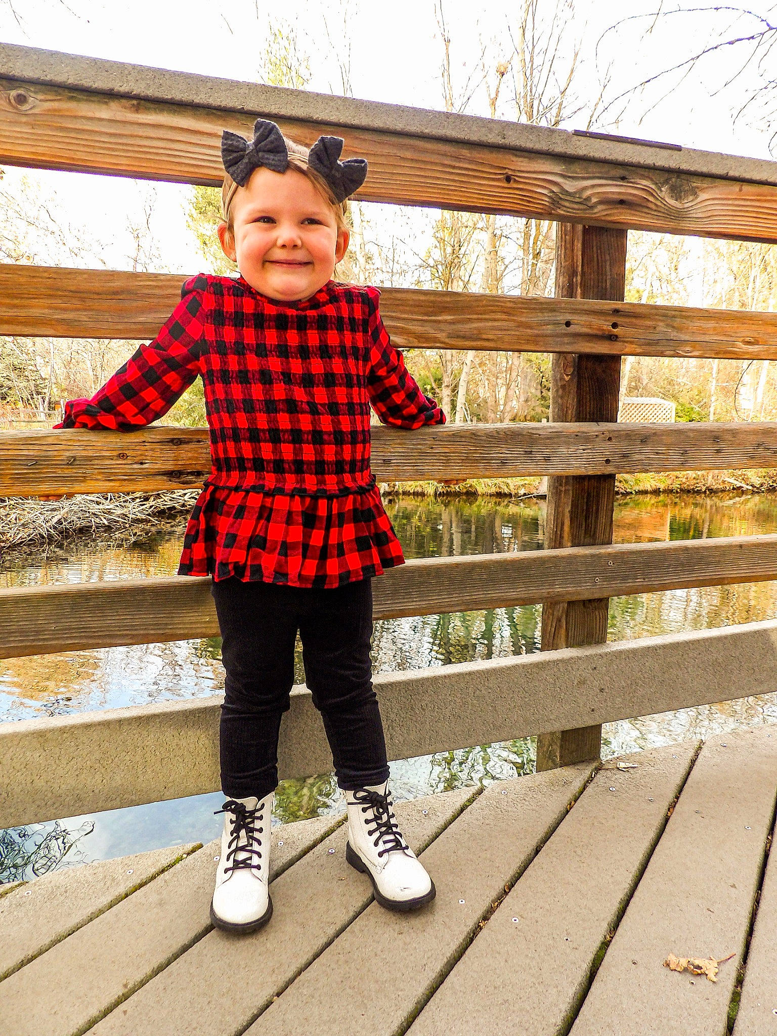 Kenleigh is registered to the contest to win money with this photo: bag, fence, footwear, grass, happy, joy, leisure, outerwear, pattern, people_in_nature, person, plaid, shoe, sleeve, sneakers, standing, street_fashion, tartan, thigh, toddler