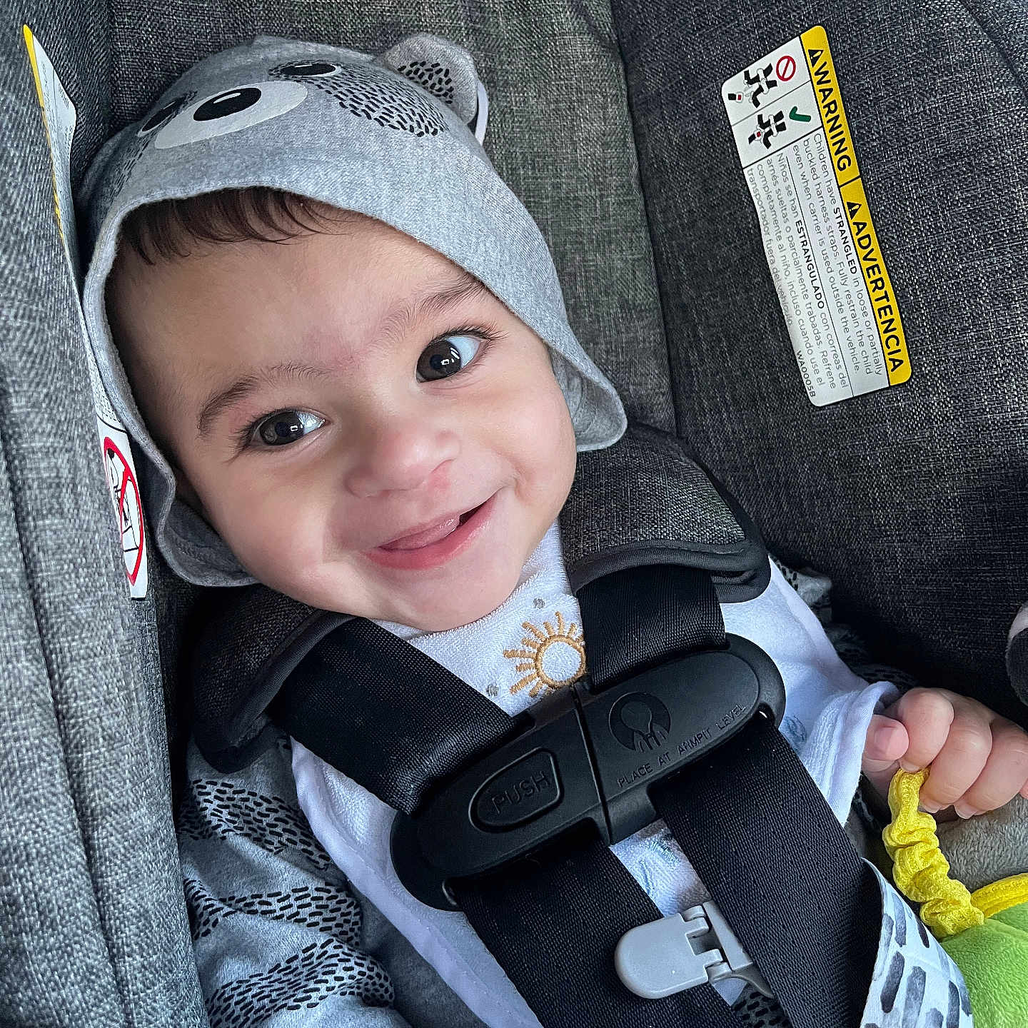 Adnan is registered to the contest to win money with this photo: baby, infant, child, smiling, happy, portrait, car_seat, seatbelt, hat, bear_hat, toy, yellow_toy, gray_upholstery, vehicle, indoors, close_up, face, eyes, white_clothing, safe
