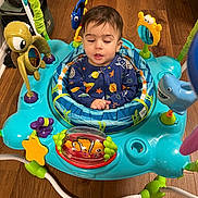 Adnan joined the competition — help win amazing prizes! baby, infant, child, sleepy, sitting, activity_walker, baby_toy, toddler, playroom, indoors, wooden_floor, colorful, animal_toy, shark_toy, turtle_toy, fish_toy, onesie, portrait, play_equipment, happy