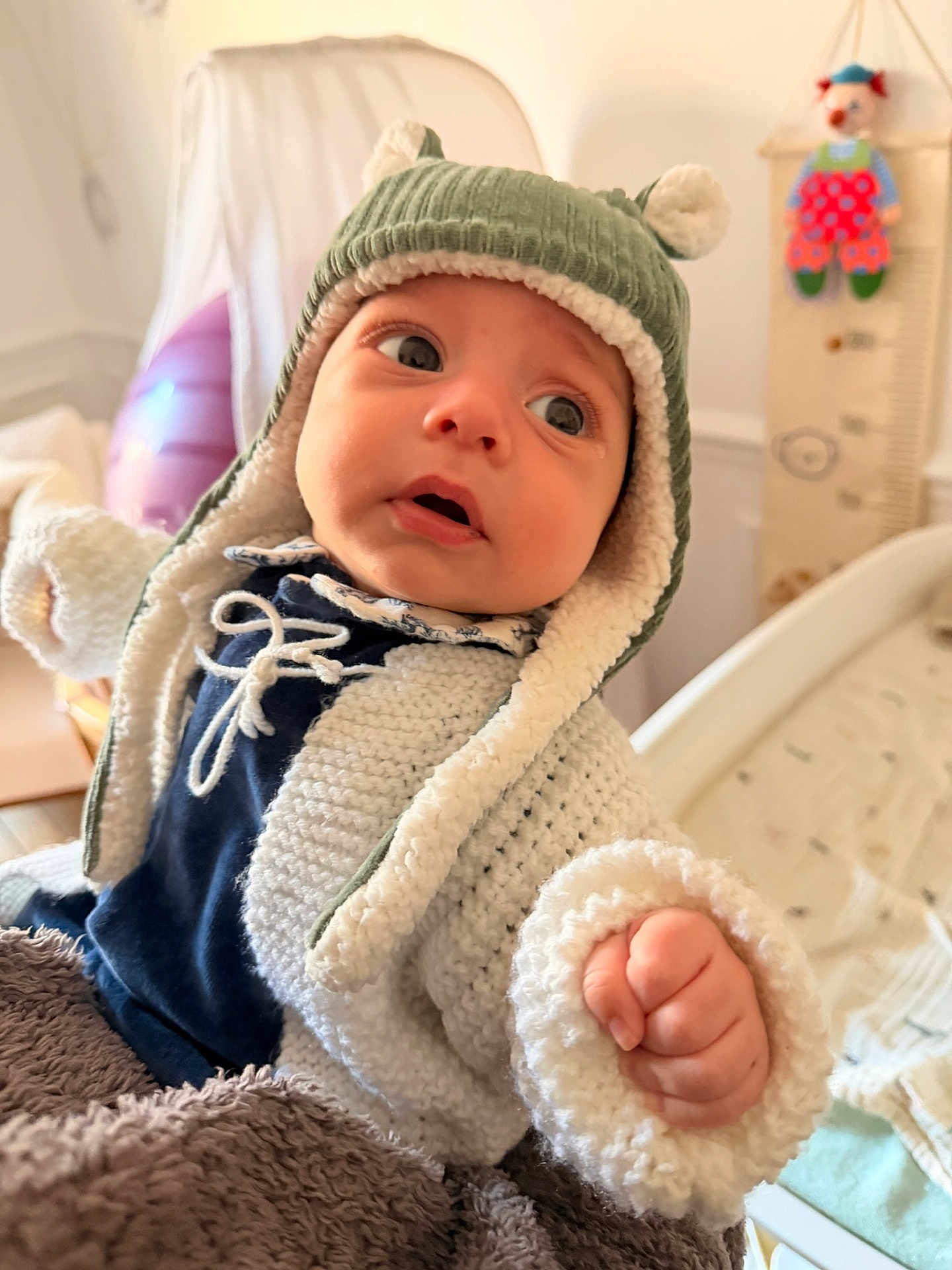 Angel a rejoint le concours — aidez-le/la à gagner de superbes lots ! baby, child, infant, knitted_jacket, hat, ear_flap_hat, blanket, indoor, portrait, face, hand, curious, soft_lighting, furniture, growth_chart, clown_toy, warm_clothing, cozy, expression, cute