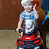 child, toddler, cap, toy_vehicle, honda, puppy, dog, indoor, floor, wooden_door, smiling, blue_eyes, striped_shirt, shorts, handlebars, pet, cute, playtime, baby, fun