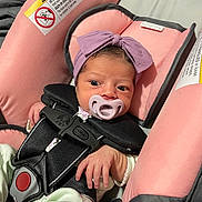 Leilani is registered to the contest to win money with this photo: baby, infant, car_seat, pacifier, headband, purple_bow, pink, clothing, straps, safety_harness, hand, face, newborn, cute, indoors, child, person, soft, comfort, portrait