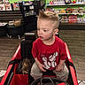 child, toy_car, red, mohawk_hairstyle, indoor, store, toys, shelves, floor, curious, clothing, tshirt, pants, seat, steering_wheel, person, young, hair, shopping, play