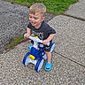 child, toddler, tricycle, outdoor, sidewalk, grass, dandelions, smile, happy, blue_sandals, shorts, shirt, toy, play, sunny_day, concrete, person, young_child, fun, recreation