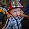 Cody joined the competition — help win amazing prizes! child, toddler, firefighter_hat, plaid_shirt, indoor, couch, adult_arm, window, casual_clothing, person, face, curious_expression, red_hat, furniture, living_room, supporting_hand, home, portrait, young_child, cozy