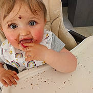 Inaya a rejoint le concours — aidez-le/la à gagner de superbes lots ! baby, child, highchair, messy, food, crumbs, face, hand, bib, eating, smiling, indoor, cute, person, infant, dining, table, cheeks, happy, tongue