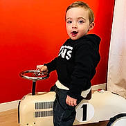 Louka participe au concours pour gagner de l'argent avec cette photo : black_hoodie, blue_eyes, child, clothing, curtain, floor, happy, indoor, light_wood_floor, number_one, orange_wall, person, playful, portrait, smiling, sneakers, steering_wheel, toddler, toy_car, vintage_toy