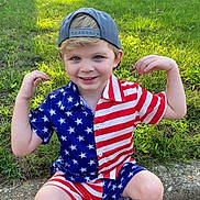 Flynn is registered to the contest to win money with this photo: child, boy, smiling, american_flag, shorts, shirt, cap, grass, lawn, curb, suburban, house, outdoor, playful, casual, footwear, sunny, daytime, pose, happy