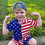 child, boy, smiling, american_flag, shorts, shirt, cap, grass, lawn, curb, suburban, house, outdoor, playful, casual, footwear, sunny, daytime, pose, happy