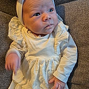Julia a rejoint le concours — aidez-le/la à gagner de superbes lots ! baby, infant, white_clothing, headband, cushion, gray_background, curious_expression, soft_lighting, small_hands, portrait, child, indoors, fabric_texture, cute, newborn, sitting, face, person, young_child, apparel
