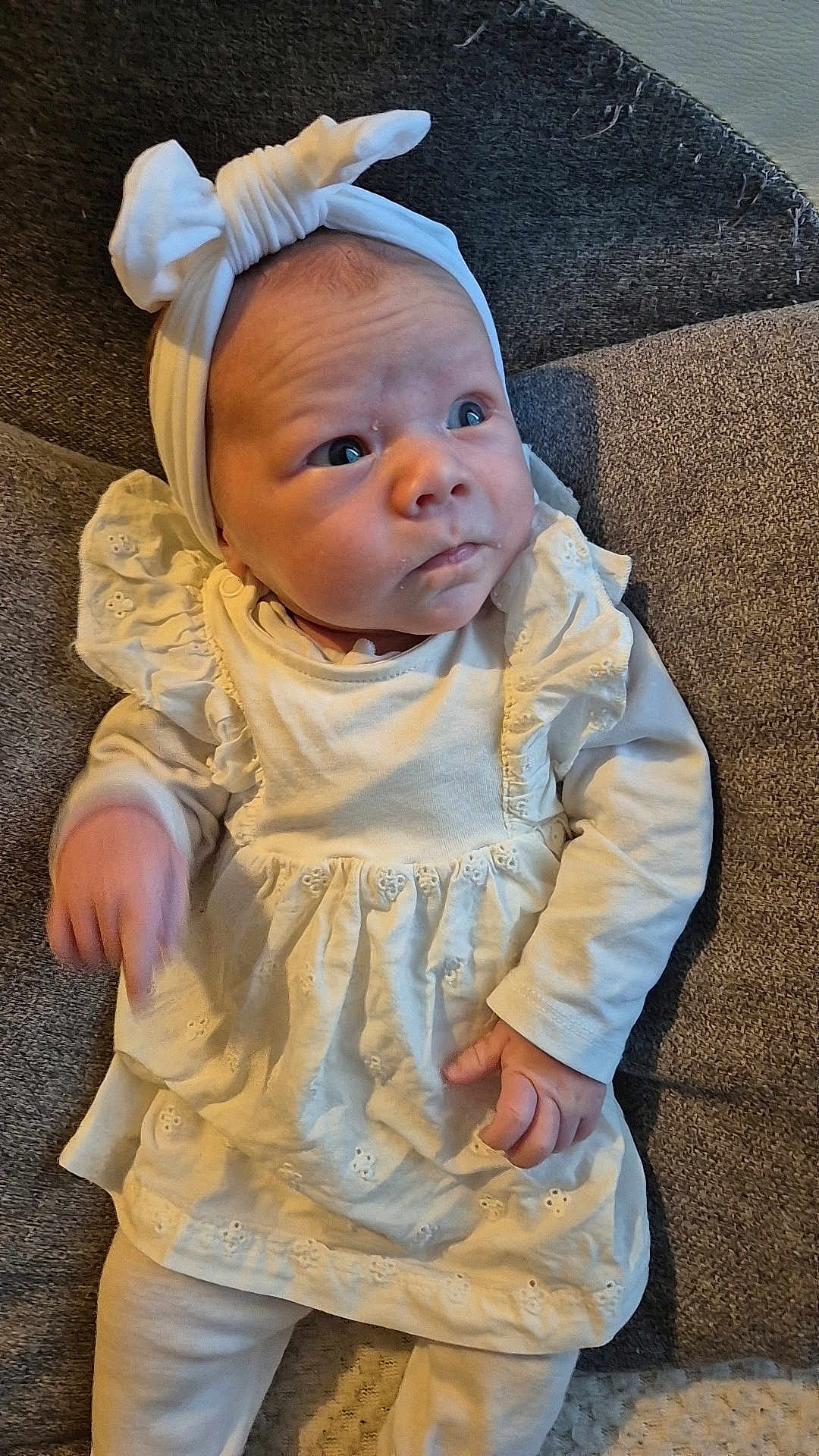 Julia a rejoint le concours — aidez-le/la à gagner de superbes lots ! baby, infant, white_clothing, headband, cushion, gray_background, curious_expression, soft_lighting, small_hands, portrait, child, indoors, fabric_texture, cute, newborn, sitting, face, person, young_child, apparel