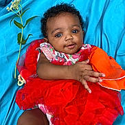 Damiyah joined the competition — help win amazing prizes! baby, child, tutu, red_dress, blue_background, flower, orange_cloth, lying_down, cute, smiling, infant, portrait, fabric, hand, face, curly_hair, bright_colors, happy, playful, soft_texture