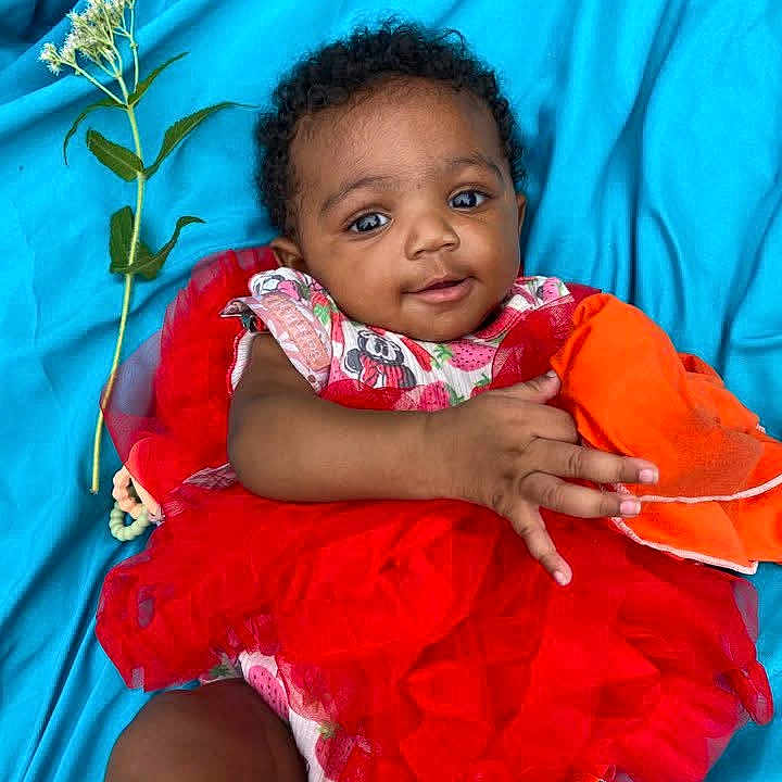 Damiyah joined the competition — help win amazing prizes! baby, blue_background, bright_colors, child, curly_hair, cute, fabric, face, flower, hand, happy, infant, lying_down, orange_cloth, playful, portrait, red_dress, smiling, soft_texture, tutu