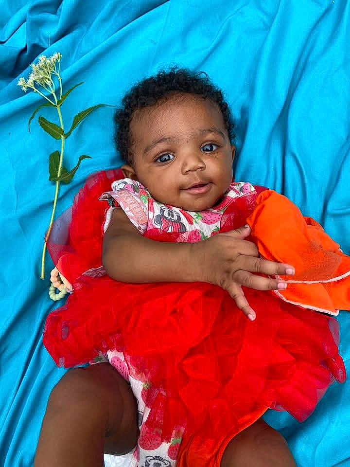 Damiyah joined the competition — help win amazing prizes! baby, child, tutu, red_dress, blue_background, flower, orange_cloth, lying_down, cute, smiling, infant, portrait, fabric, hand, face, curly_hair, bright_colors, happy, playful, soft_texture