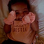 baby, infant, smiling, pink_mittens, shirt, text_on_clothing, pillow, blanket, lying_down, cute, child, indoor, cozy, happy, face, hands, clothing, soft, bed, resting