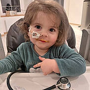 Nina participe au concours pour gagner de l'argent avec cette photo : baby, child, curly_hair, cute, face, feeding_tube, green_clothing, healthcare, high_chair, home, indoor, kitchen, medical, nasal_tube, person, smile, stethoscope, table, toddler, window