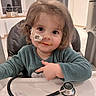 baby, child, curly_hair, cute, face, feeding_tube, green_clothing, healthcare, high_chair, home, indoor, kitchen, medical, nasal_tube, person, smile, stethoscope, table, toddler, window