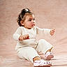 baby, child, cream_color, curly_hair, cute, earrings, expression, fashion, footwear, hair_accessory, indoor, knitwear, person, pink_shoes, portrait, sitting, soft_background, studio, toddler, young_child