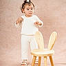 Everly is registered to the contest to win money with this photo: beige_background, bunny_ears, child, cream_color, curly_hair, cute, expression, footwear, furniture, indoor, knitwear, minimalist, playful, portrait, sandals, standing, studio, toddler, wooden_chair, young_child