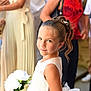 child, girl, flower_bouquet, white_dress, hair_accessory, smile, portrait, event, formal_clothing, people, crowd, blurred_background, standing, happy, outdoor, celebration, fashion, person, young, cute