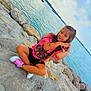 child, girl, smiling, pink_shirt, black_shorts, sneakers, rocks, water, sea, sky, outdoor, sitting, long_hair, casual_clothing, playful, nature, coast, daylight, portrait, happy