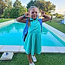 child, girl, smiling, playful, turquoise_dress, white_sneakers, backpack, pool, grass, outdoor, trees, daylight, pose, happy, young, recreation, casual_clothing, nature, blue_sky, person